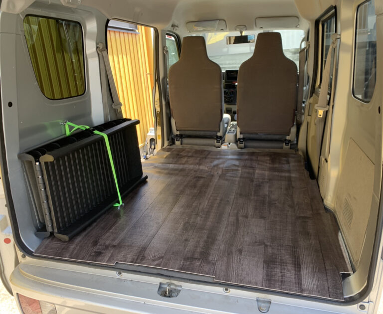 後部ドアから見た軽バンの荷室。木目調の床材を敷き、折りたたんだペット用ケージを左の側面にベルトで固定している